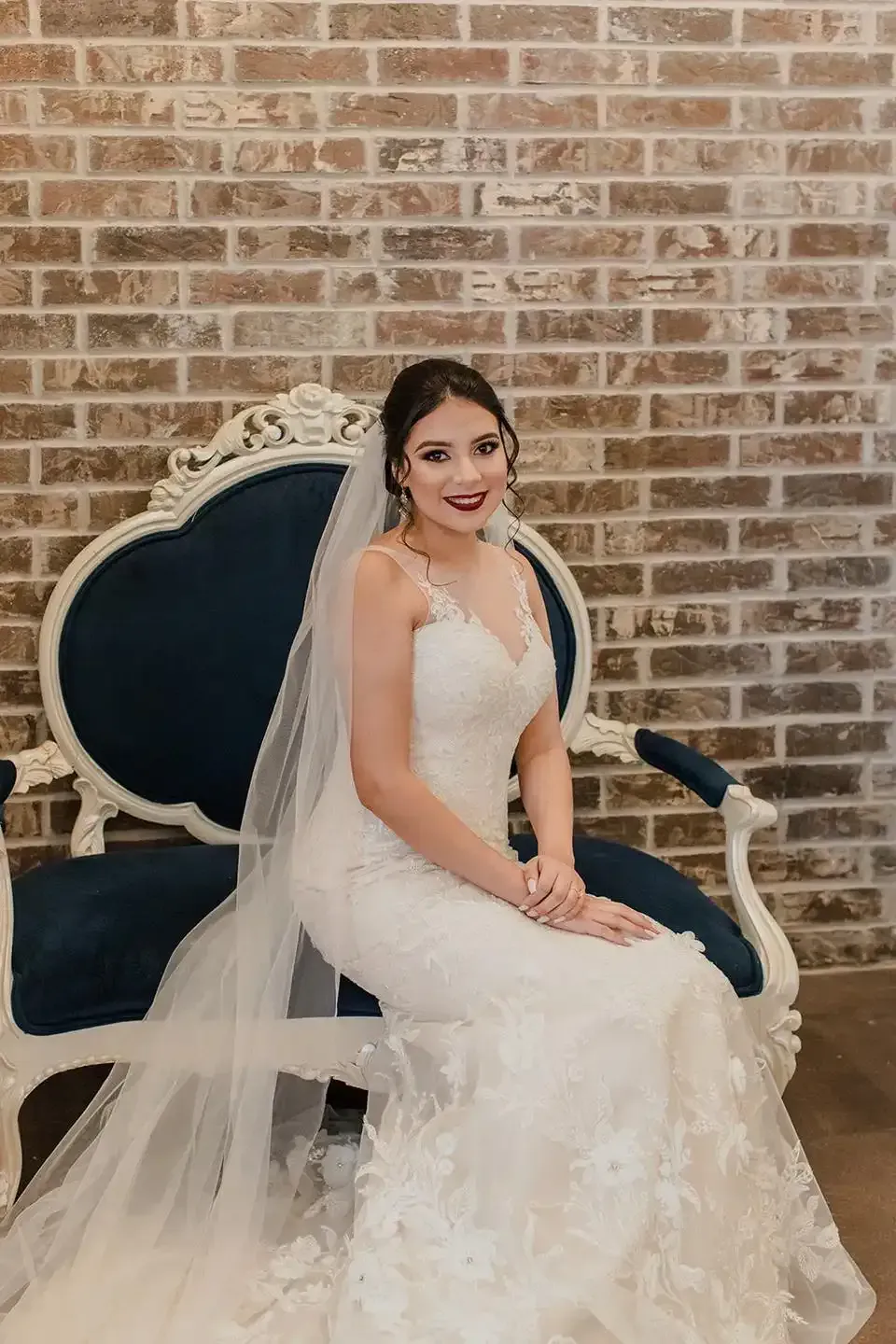 A bride in a wedding dress is sitting on a chair in front of a brick wall.