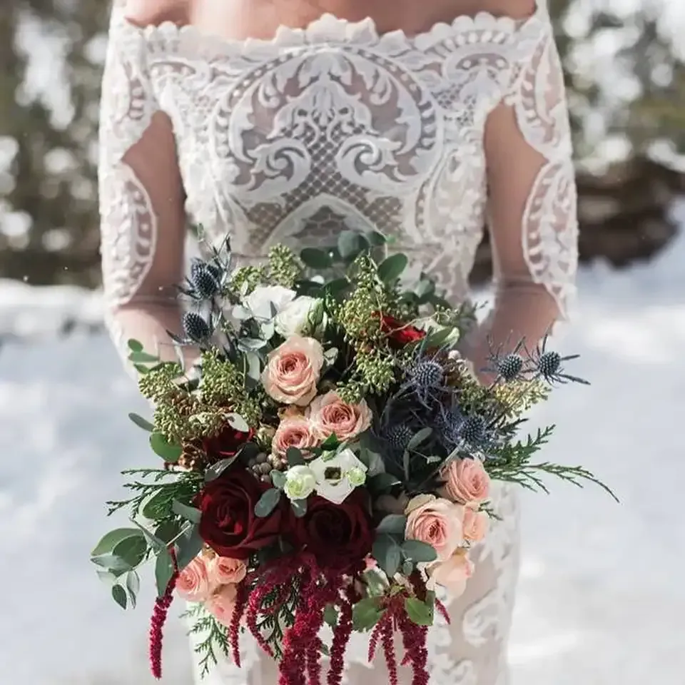 A bride in a white lace dress is holding a bouquet of flowers in the snow.