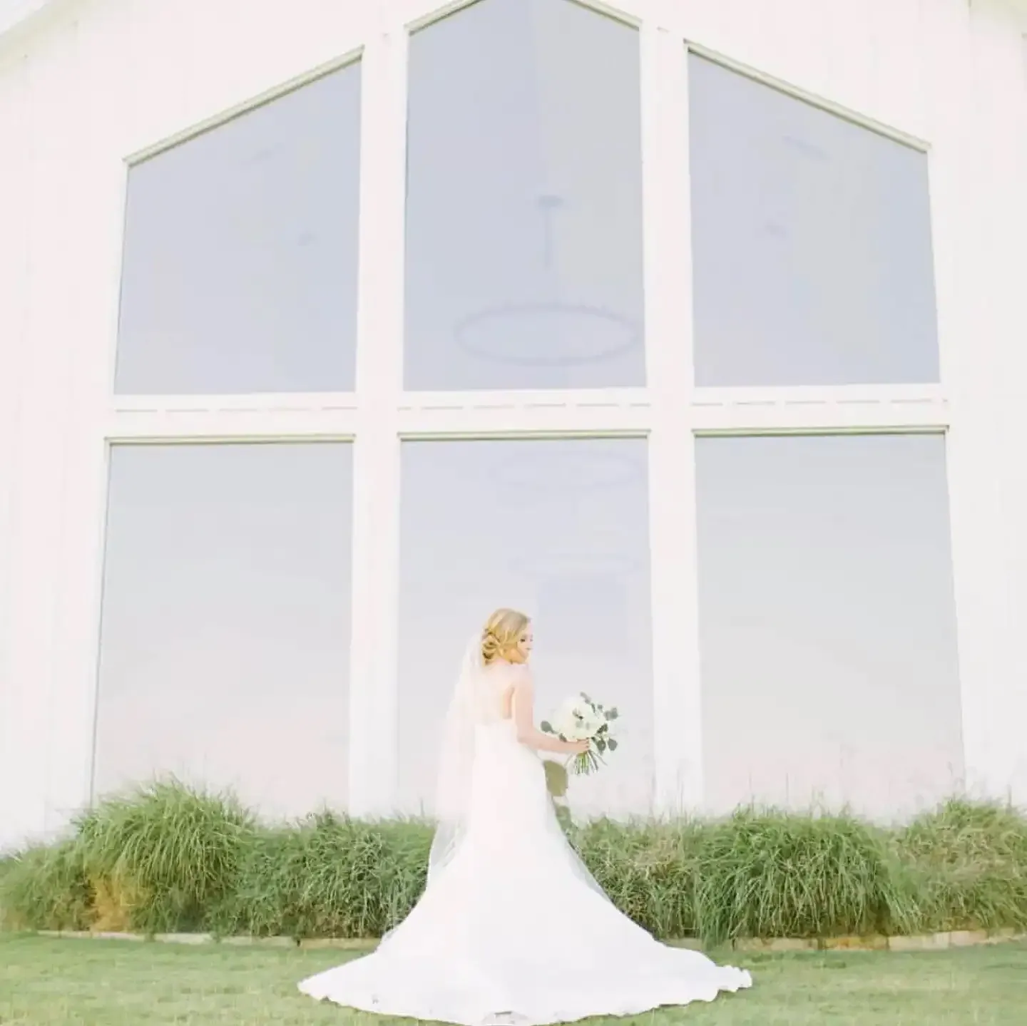 A bride in a wedding dress and veil is standing in front of a white building holding a bouquet of flowers.