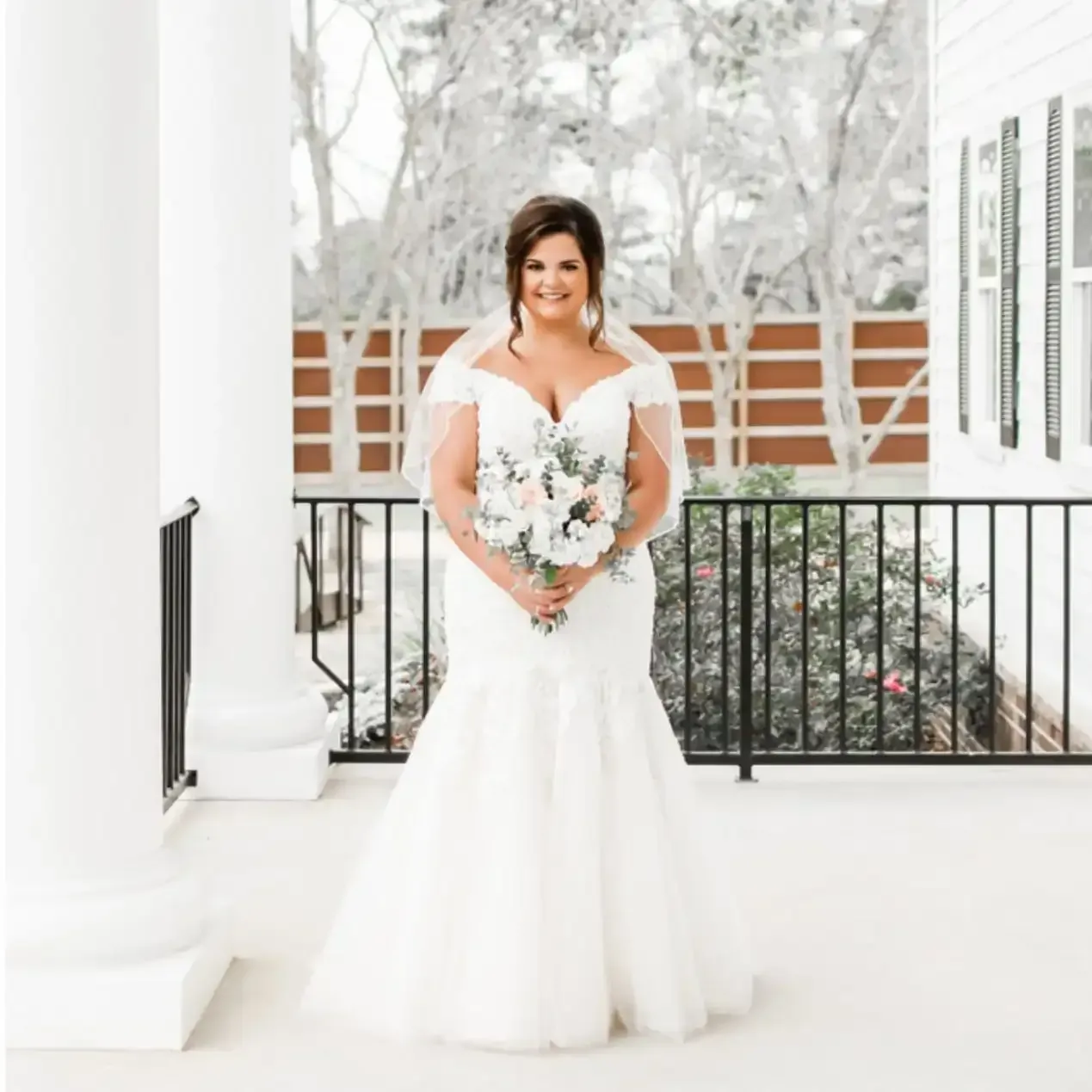 A bride in a wedding dress is standing on a porch holding a bouquet of flowers.