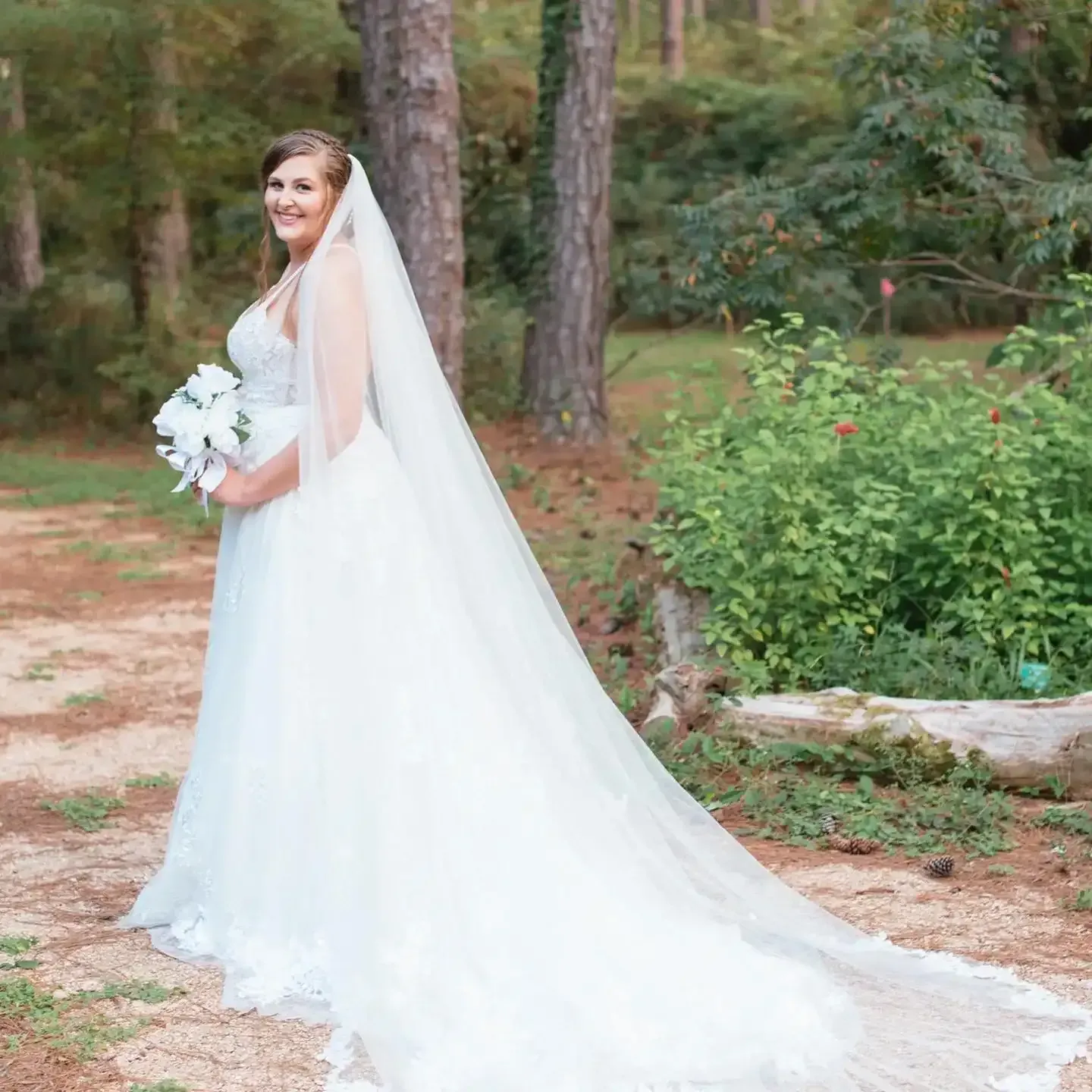 A bride in a white wedding dress and veil is standing in the woods holding a bouquet of flowers.