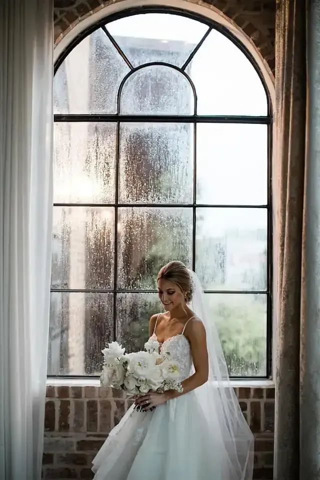 A bride in a wedding dress is standing in front of a window holding a bouquet of flowers.