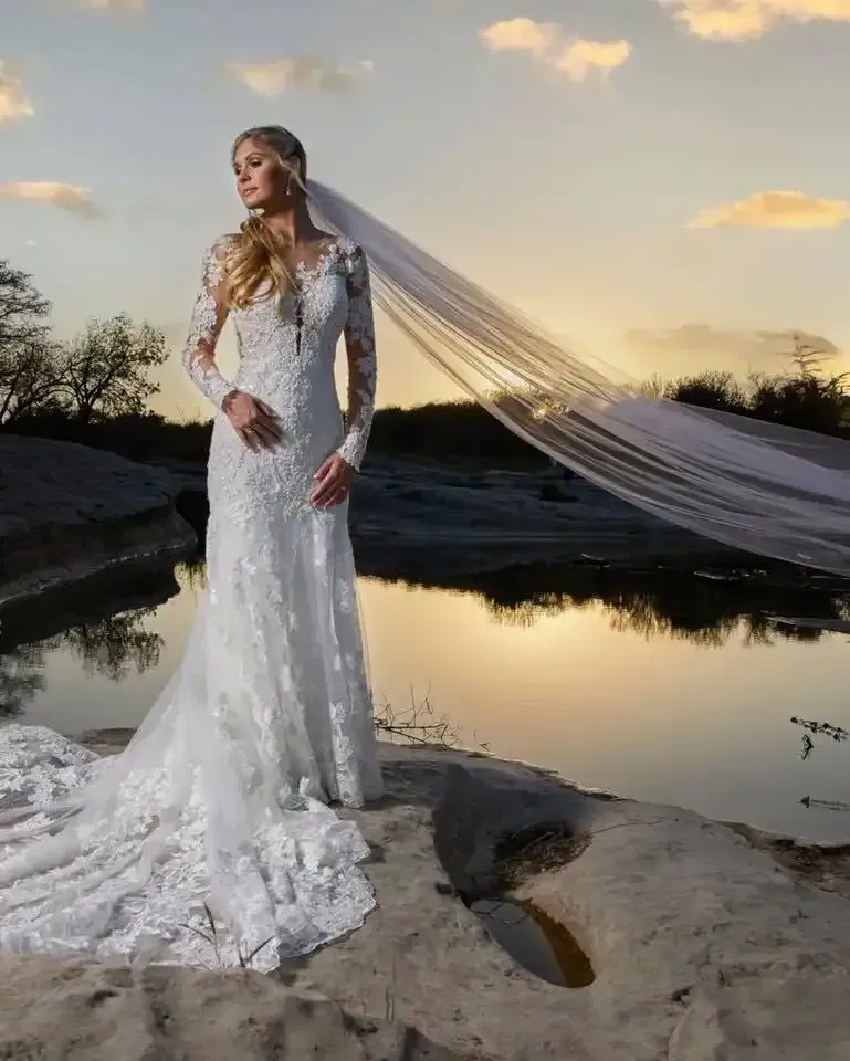 A woman in a wedding dress and veil is standing in front of a body of water.