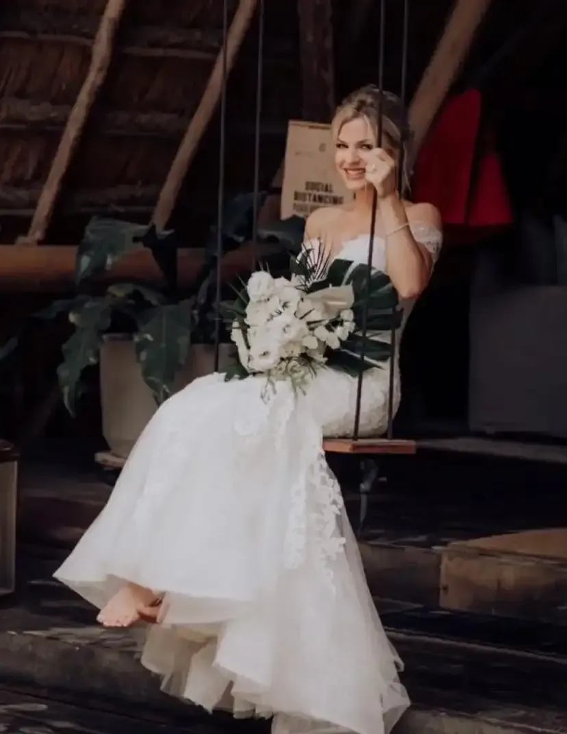 A woman in a wedding dress is sitting on a swing holding a bouquet of flowers.