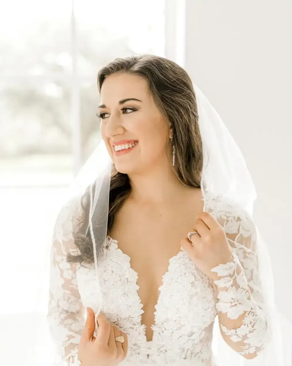 The bride is wearing a veil and smiling while standing in front of a window.