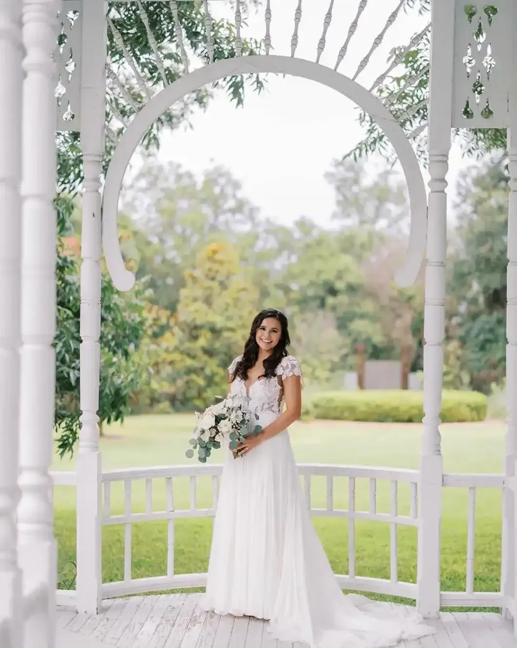 A bride in a white dress is standing in a gazebo holding a bouquet of flowers.