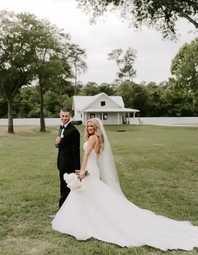 A bride and groom are standing in a grassy field in front of a white house.