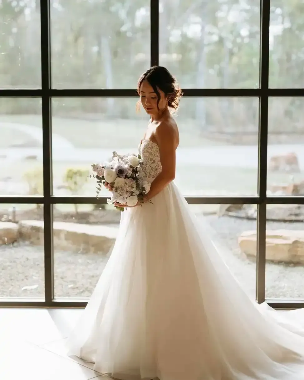 A bride in a white wedding dress is standing in front of a window holding a bouquet of flowers.