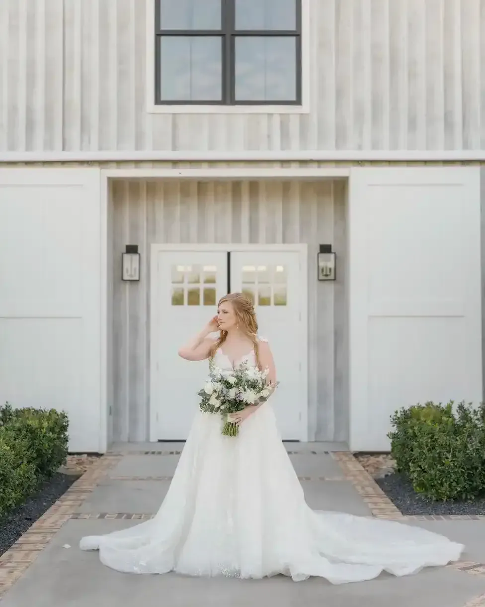 A bride in a wedding dress is standing in front of a white building holding a bouquet of flowers.