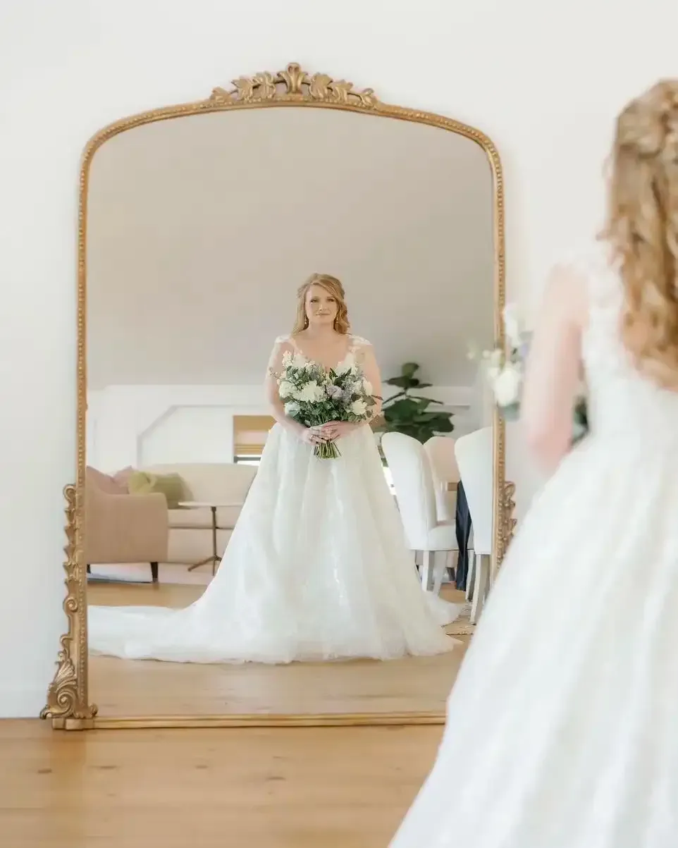 A bride is standing in front of a large mirror holding a bouquet of flowers.
