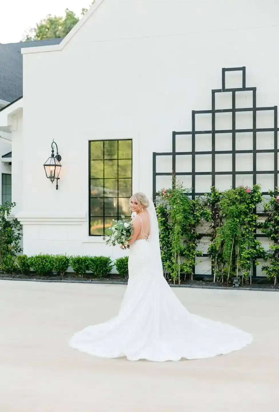 A bride in a wedding dress is standing in front of a white building holding a bouquet of flowers.