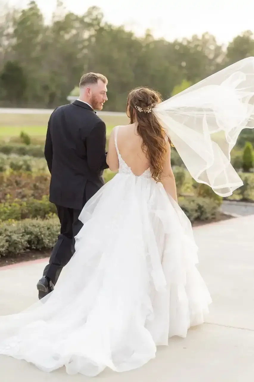 A bride and groom are walking down a sidewalk with their veil blowing in the wind.
