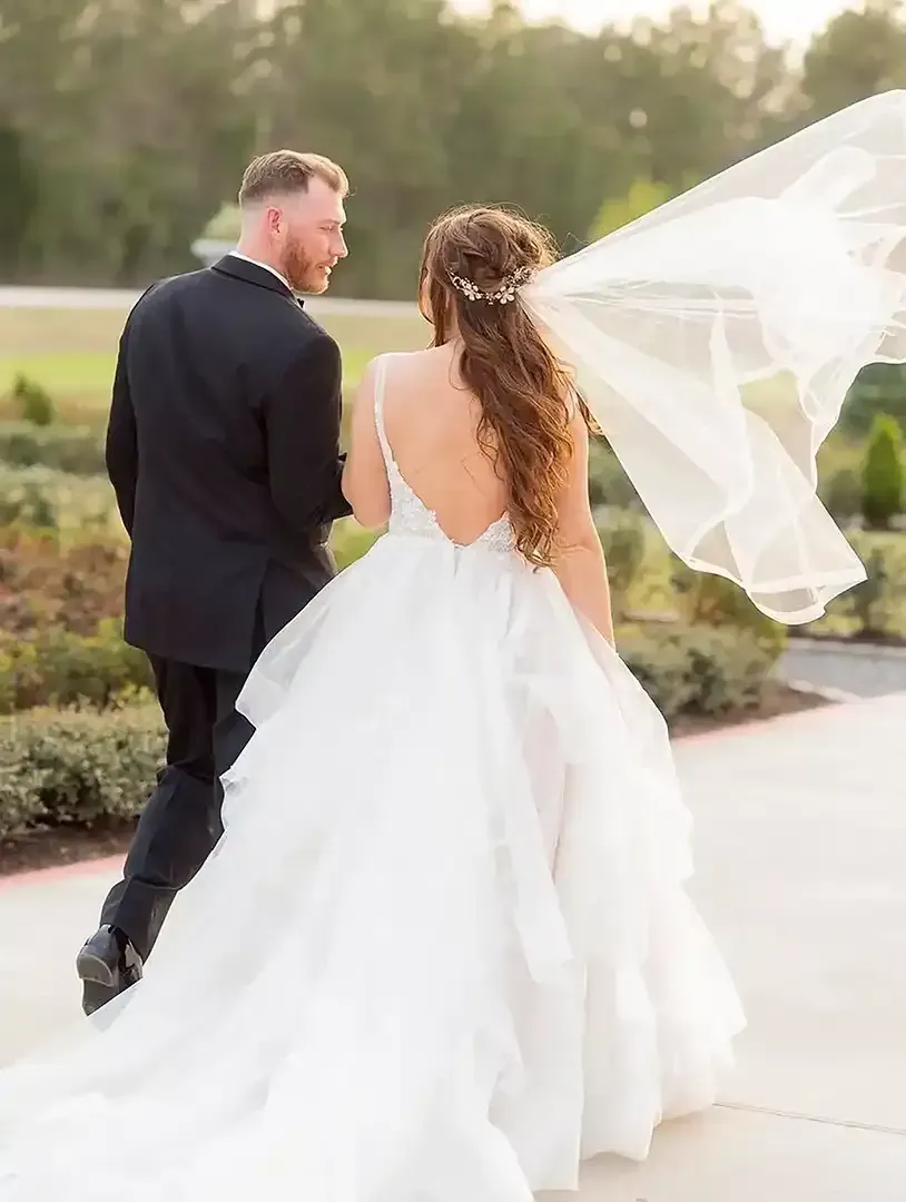 A bride and groom are walking down a sidewalk with their veil blowing in the wind.