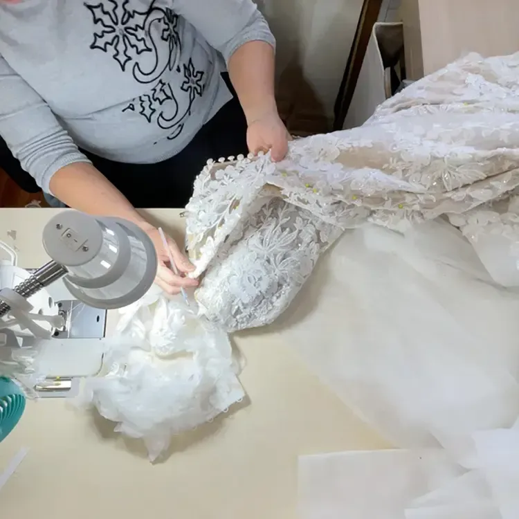 Person sewing a white lace wedding dress at a table. Luxury Tailors by Monica