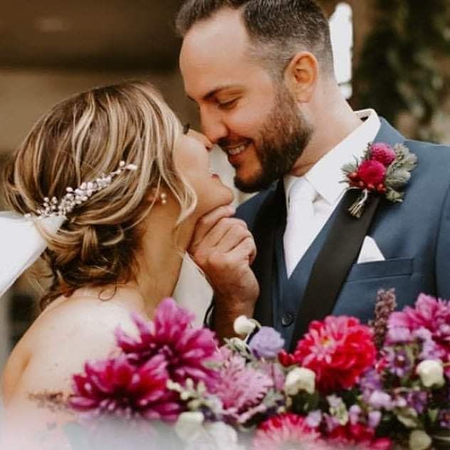 Bride and groom embrace, close-up. She has a flower crown, he wears a blue suit and smiles. Purple flowers in foreground.