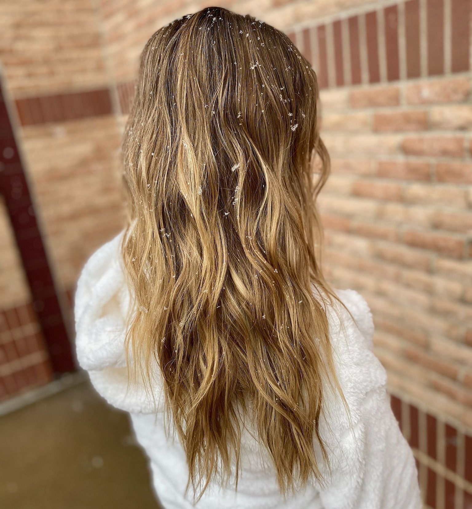 Woman with long, wavy, blonde hair wearing a white hooded robe, standing in front of a brick wall.