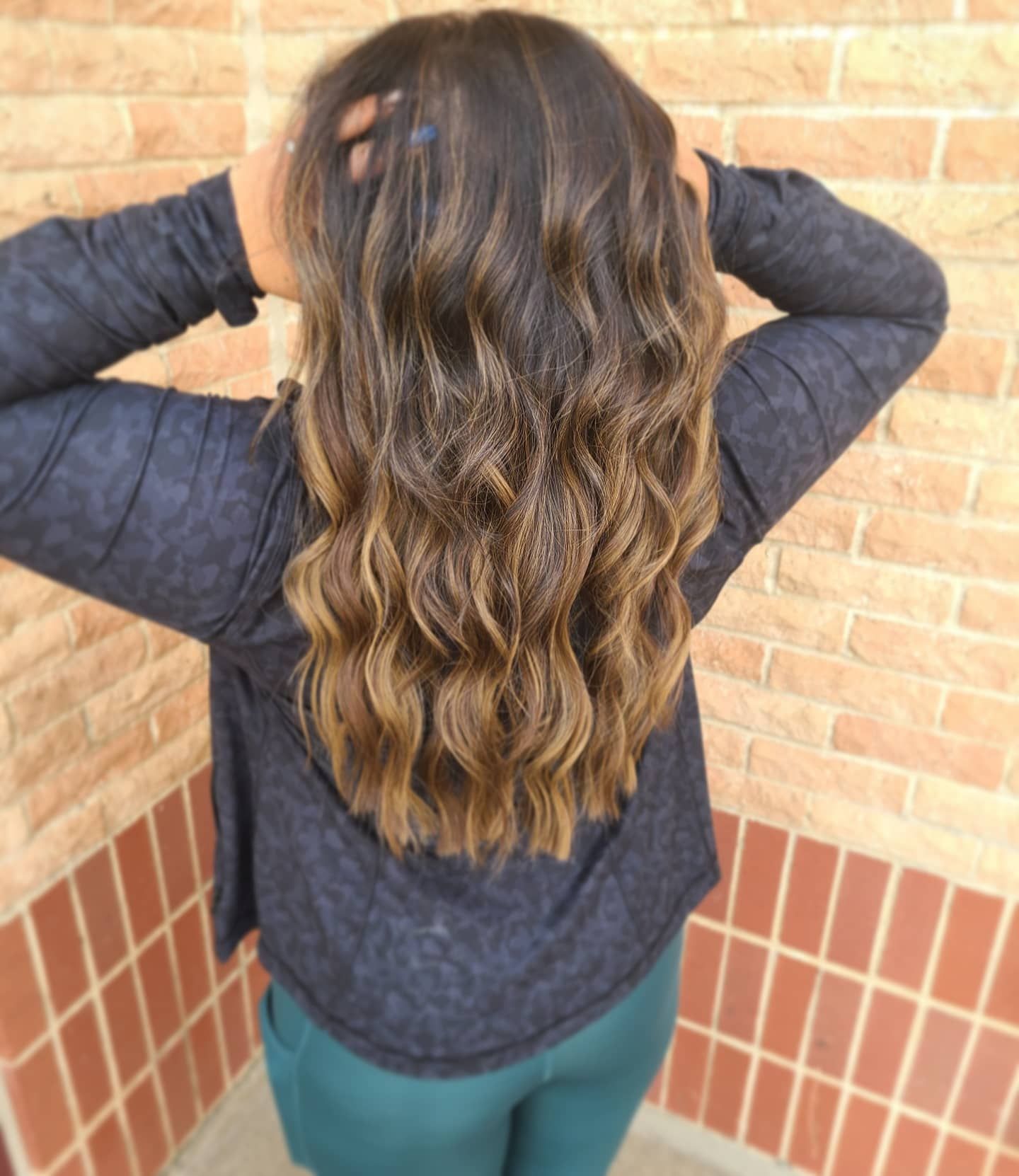 Woman with wavy brown hair, hands behind head. Hair is highlighted, wearing a dark shirt and teal pants. Brick wall background.