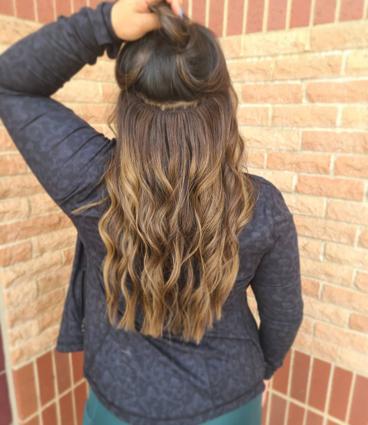 Woman with wavy, ombre hair, in front of a brick wall. Her hair is partly up, wearing a dark shirt.