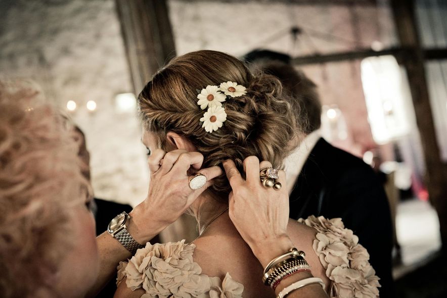 Woman's hair styled with flowers, being adjusted by hands wearing jewelry, in a dim setting.