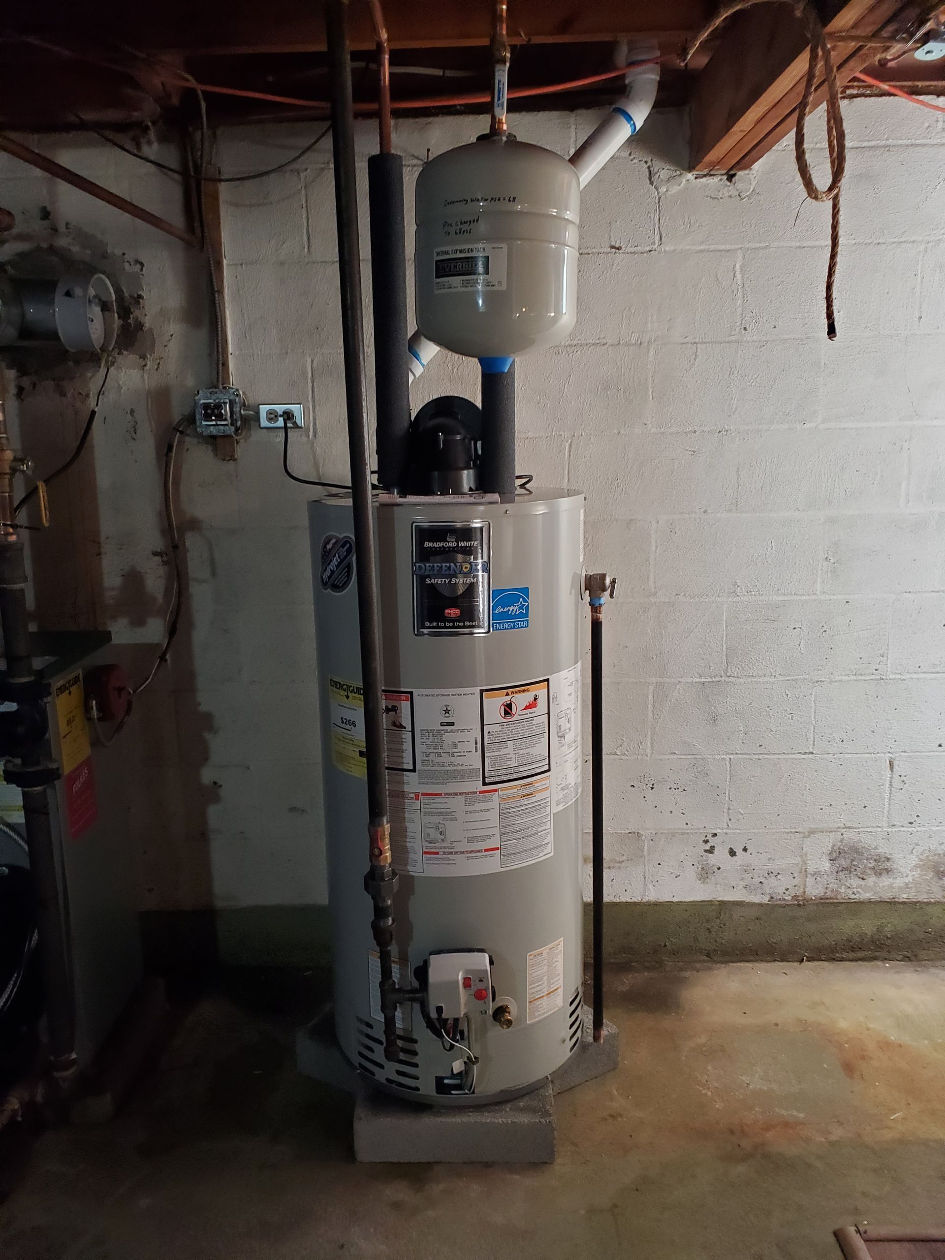A water heater is sitting in a basement next to a brick wall.