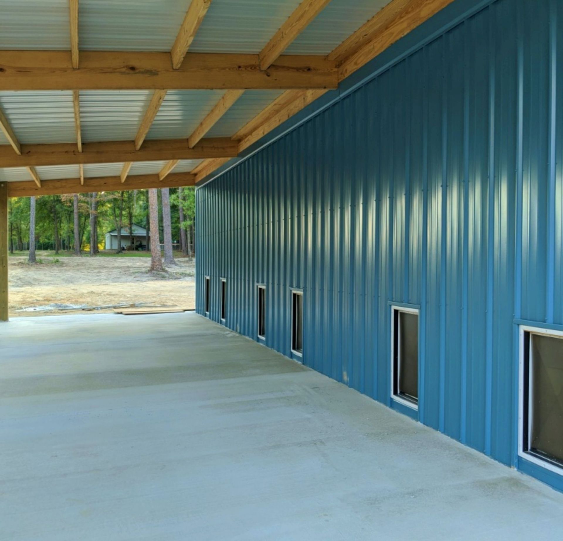 Blue metal building with small windows beside a concrete covered walkway and wooden roof beams