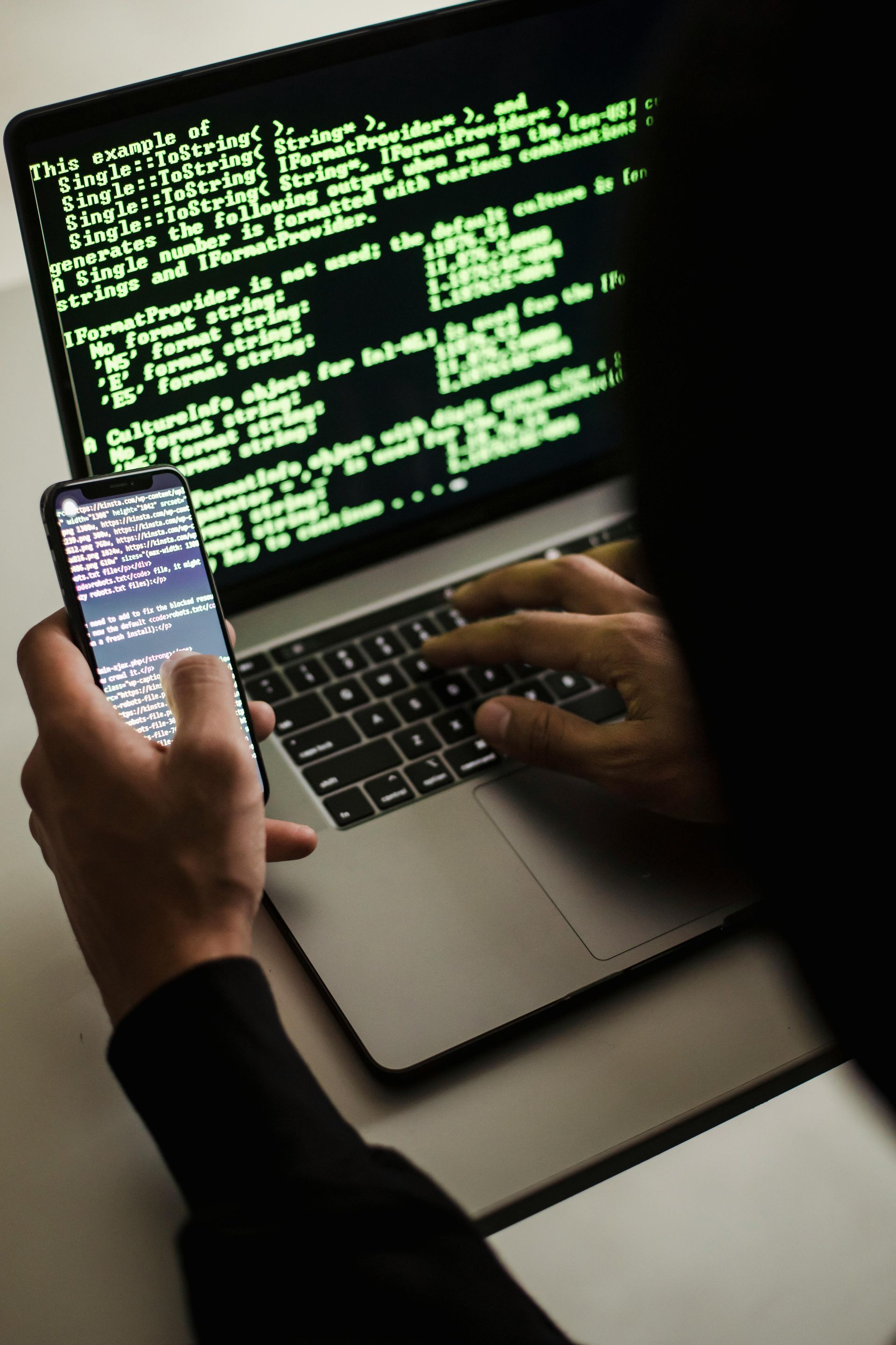 A person is typing on a laptop while holding a cell phone