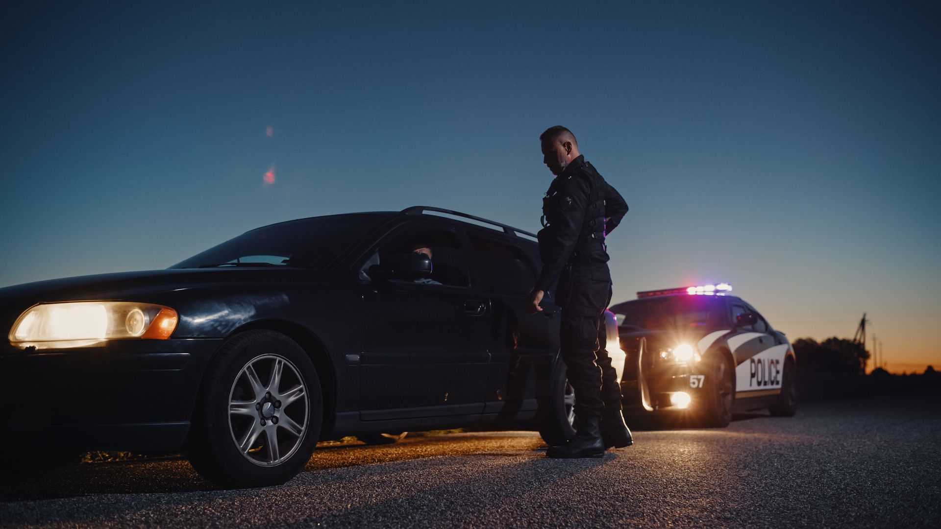 A police officer stands beside a dark car pulled over at night, patrol car lights behind. A police officer stands beside a dark car pulled over at night, patrol car lights behind.