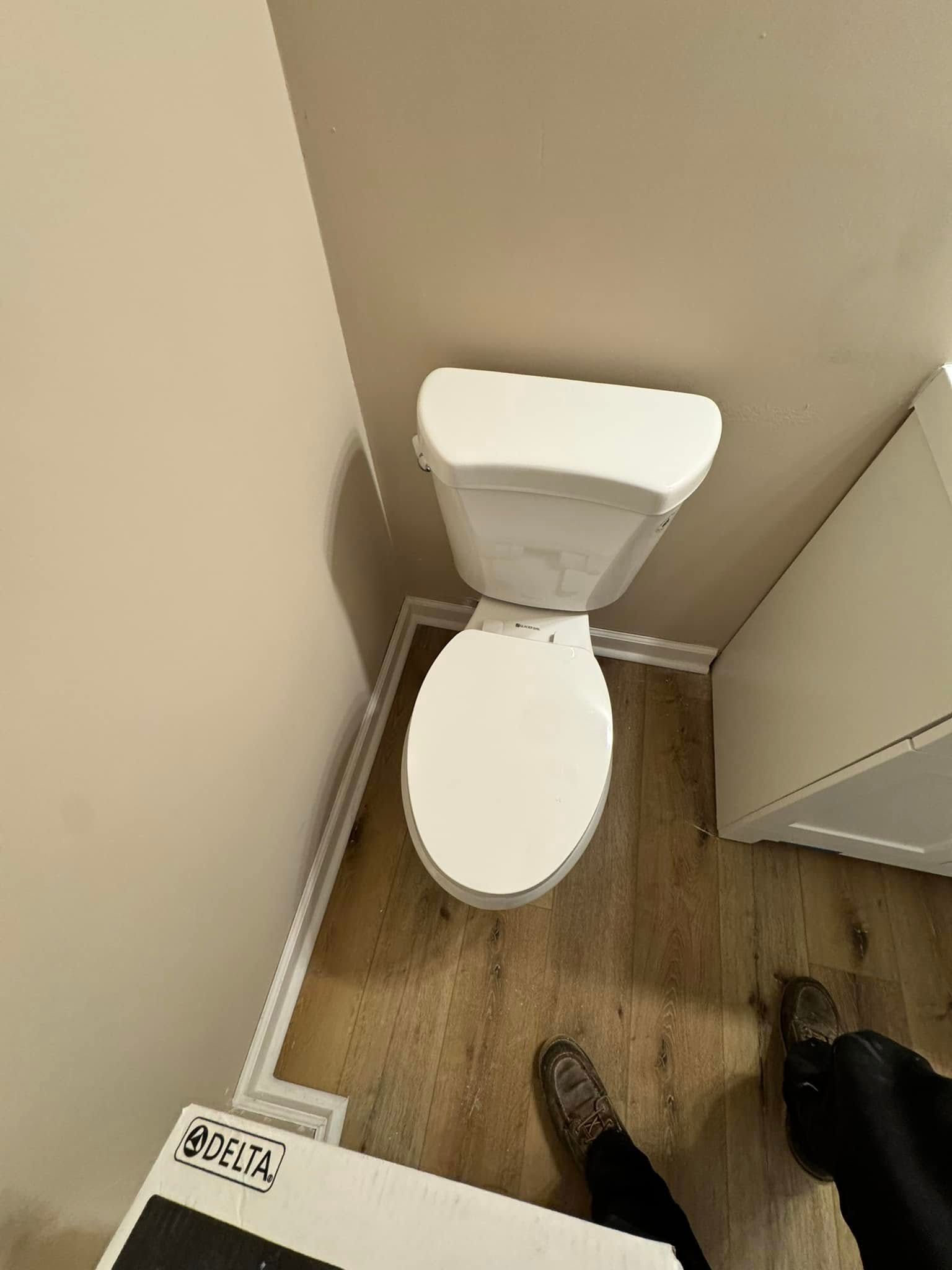 Toilet in a bathroom with wood-look flooring and a small cabinet. A person's feet are visible.