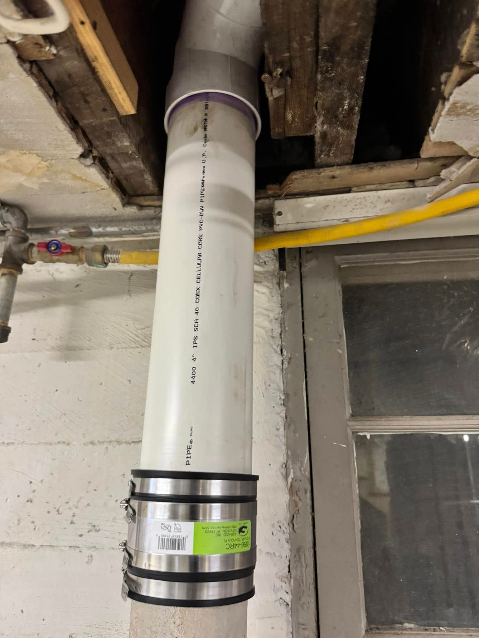 White PVC pipe extending through a ceiling, connected with metal clamps. Wooden beams and a window are in the background.