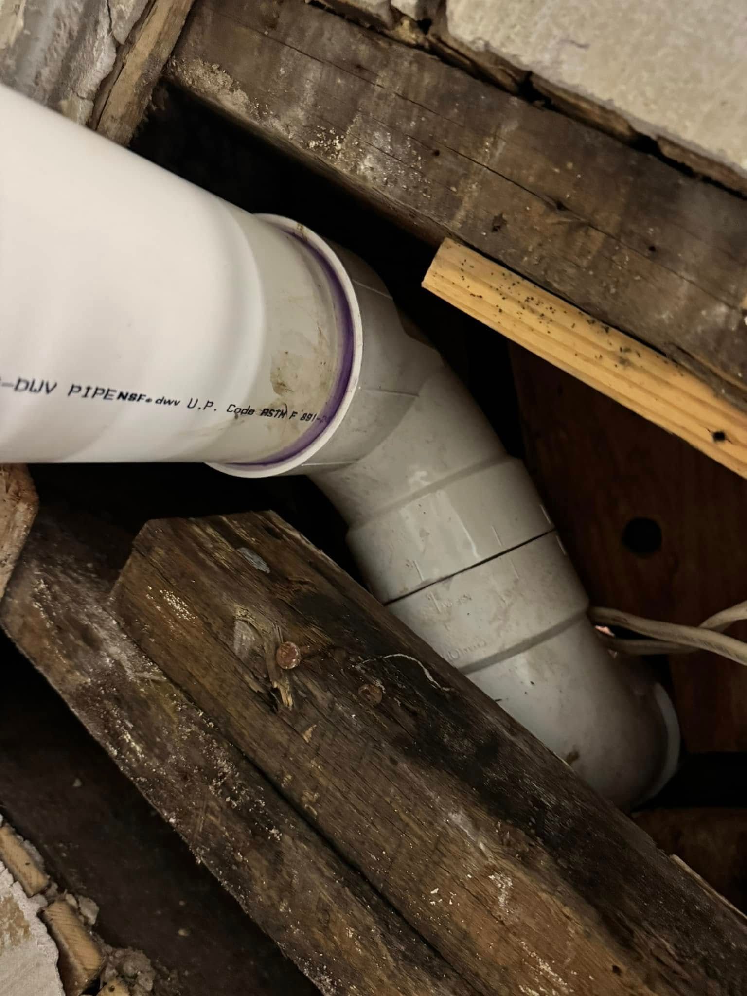 White PVC pipe with a 90-degree elbow connected to another section, surrounded by wooden beams and dark debris.