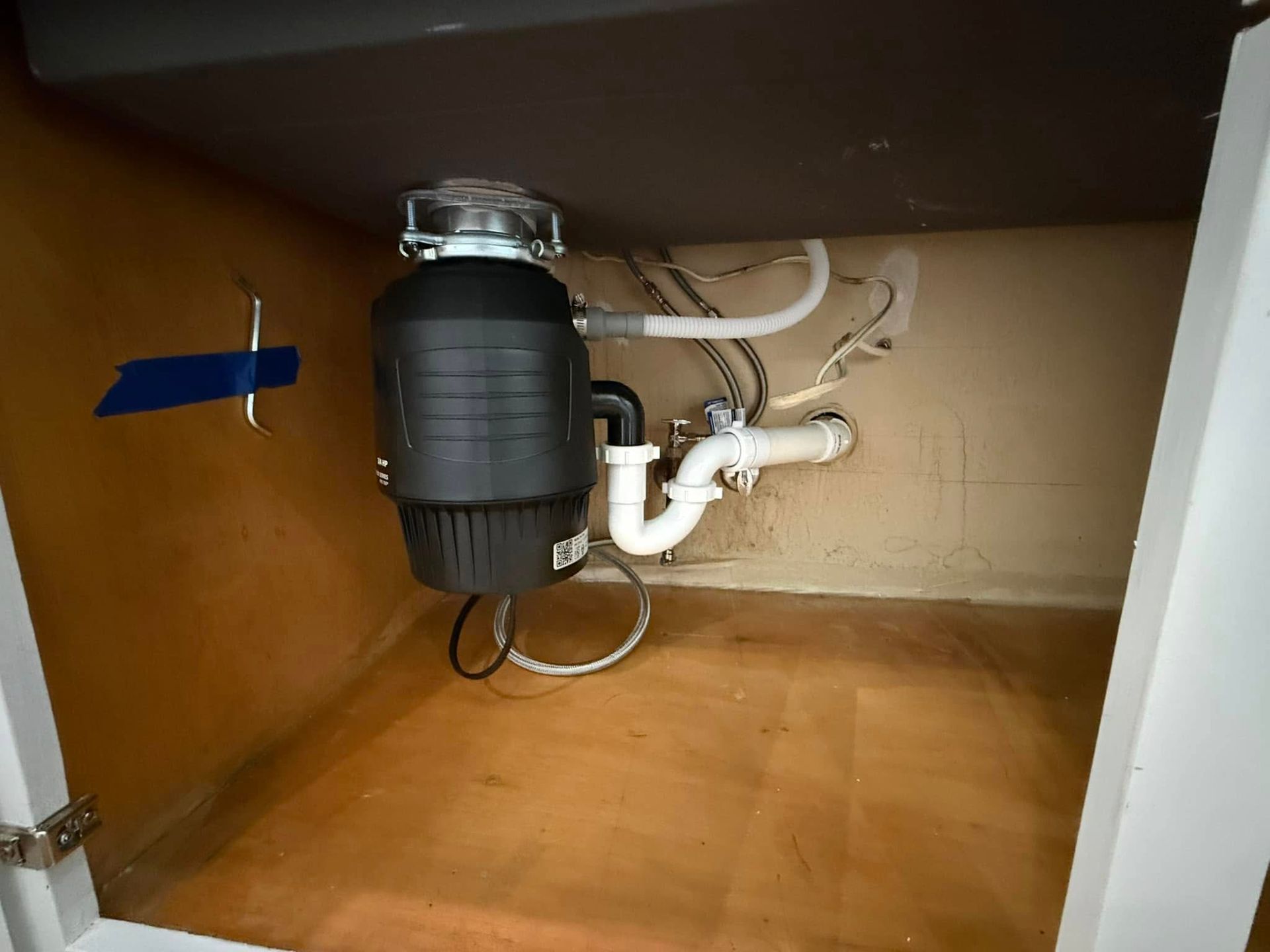 Garbage disposal plumbing under a kitchen sink. White pipes and disposal against beige cabinet.
