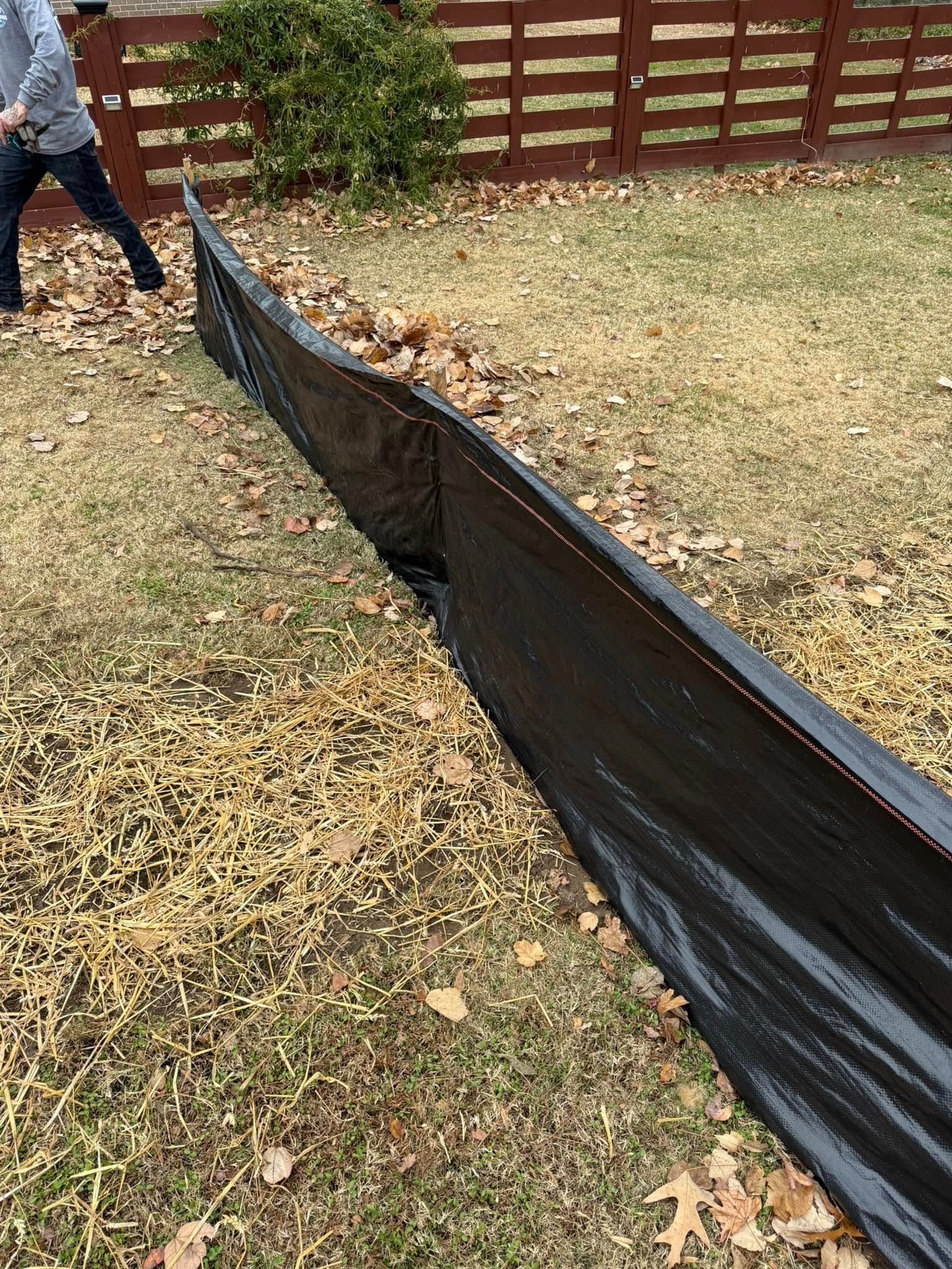 Black tarp containing leaves on a grassy lawn, with a wooden fence in the background. A person stands nearby.