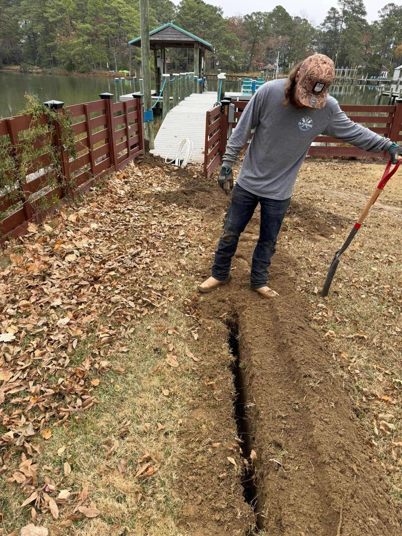 Man digging a trench in a yard near a lake with a shovel. Brown and green landscape.