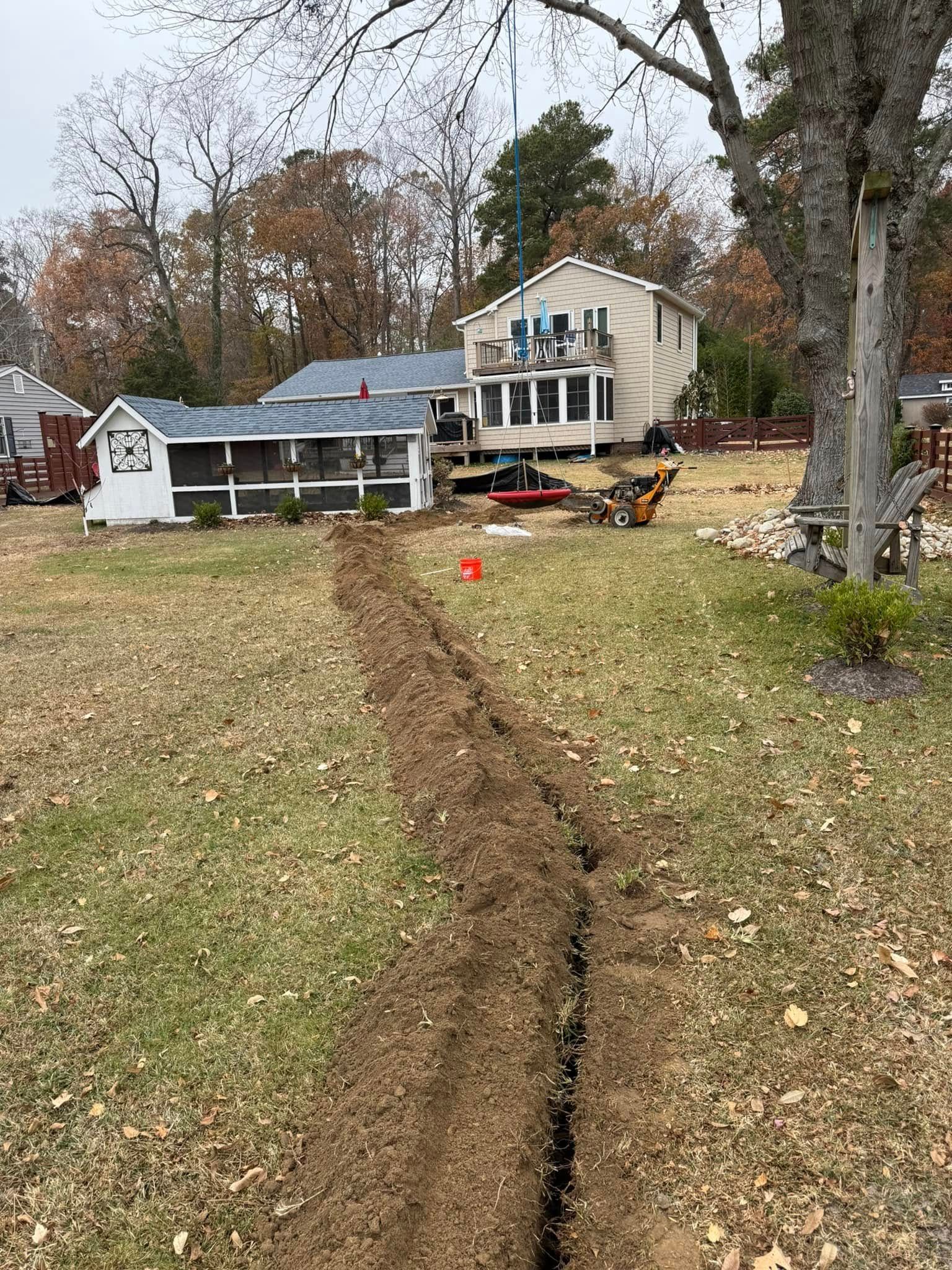 Trench dug in a grassy yard, leading toward a house. A small shed and equipment are nearby. Cloudy day.