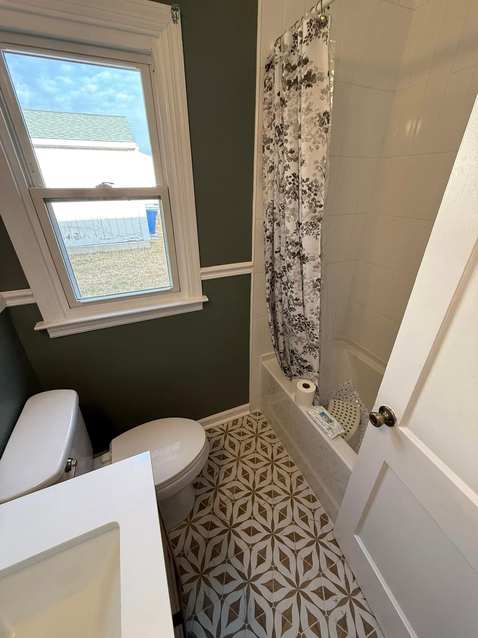 Bathroom with olive green walls, patterned floor, toilet, bathtub, and window.