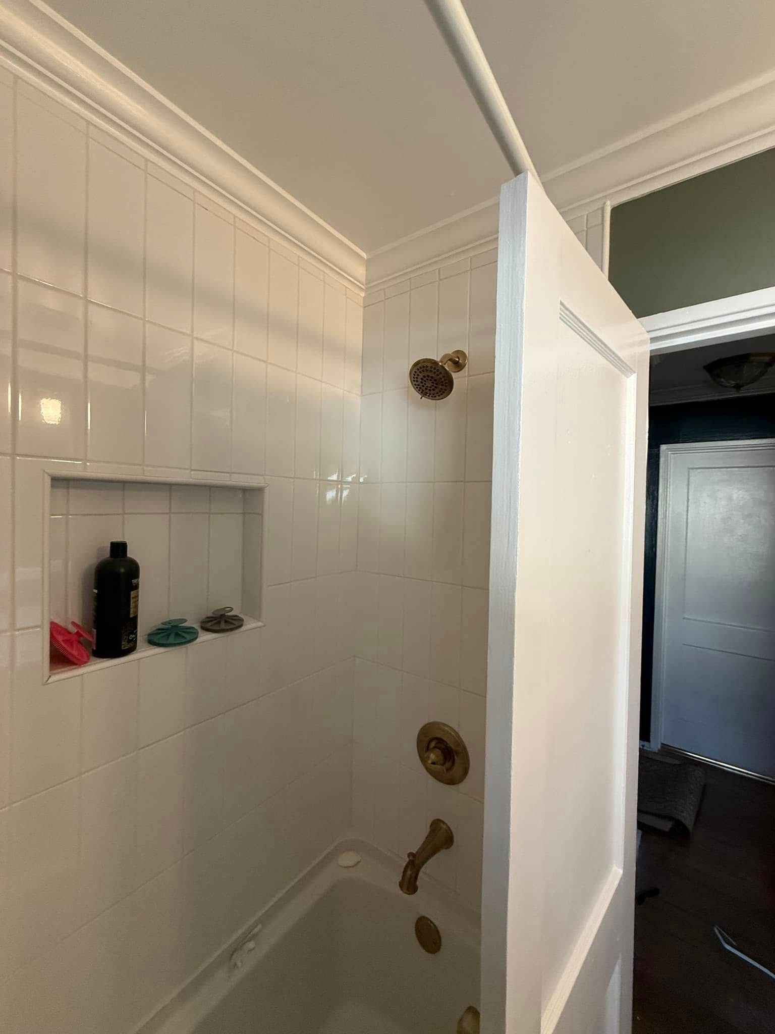 White-tiled shower with gold fixtures, recessed shelf, and partial view of a white door and adjacent room.