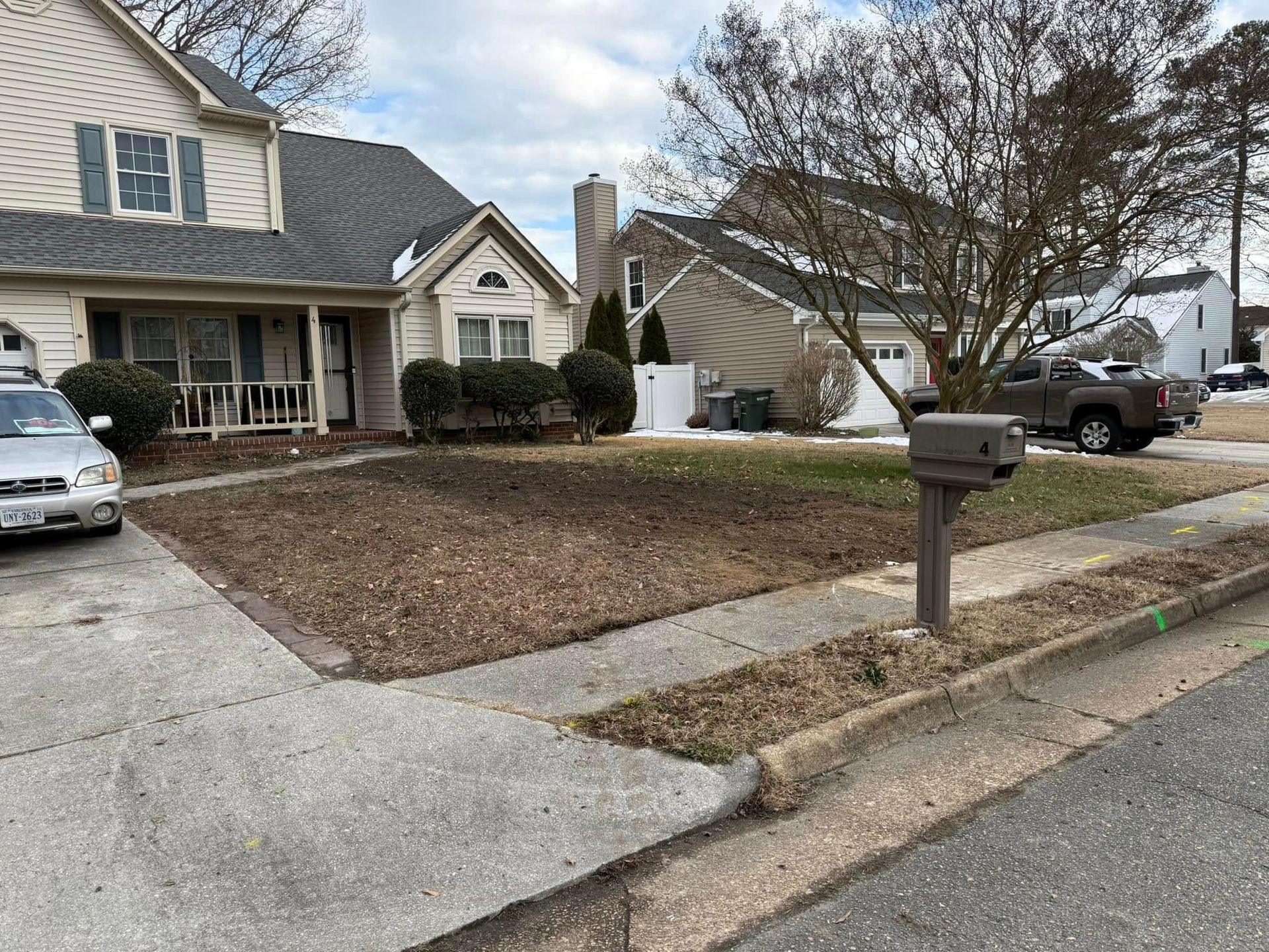 House with bare front yard; car parked on left, mailbox in the foreground, and a truck parked in the right background.