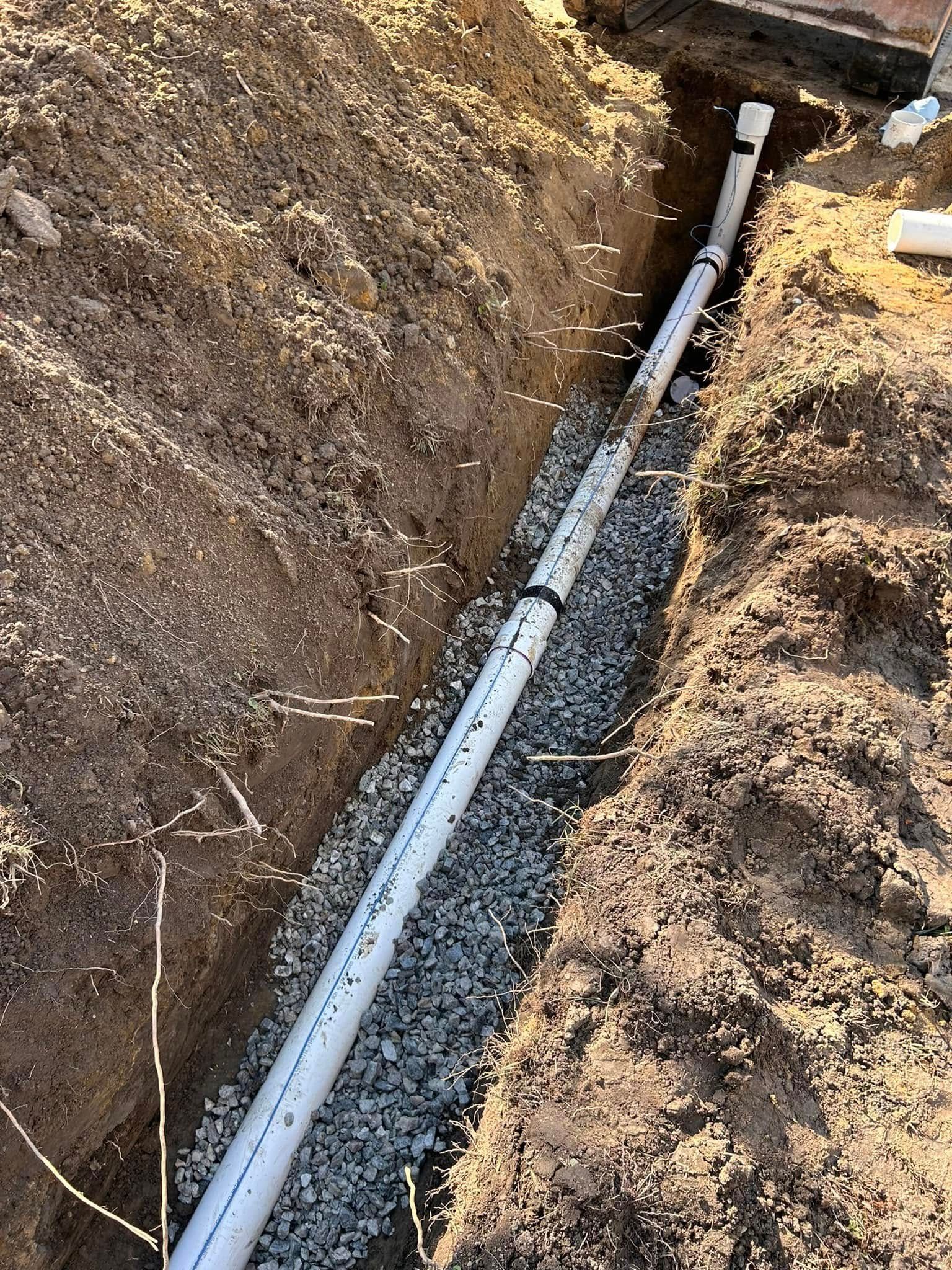 A PVC pipe installed in a trench lined with gravel. Dirt and grass surround the trench.