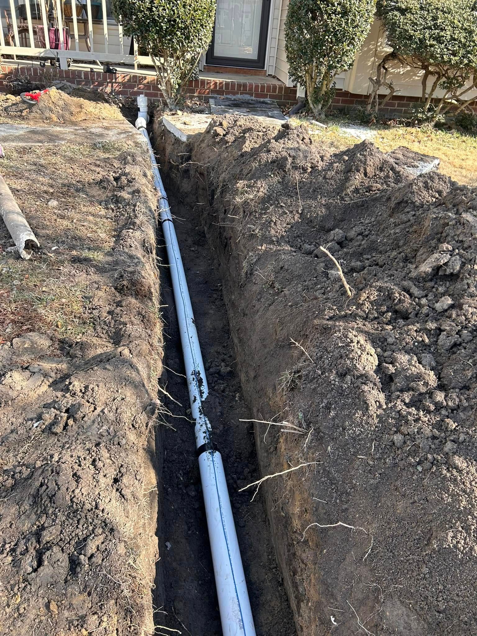 Trench with white PVC pipe installed, running along a walkway in front of a house.