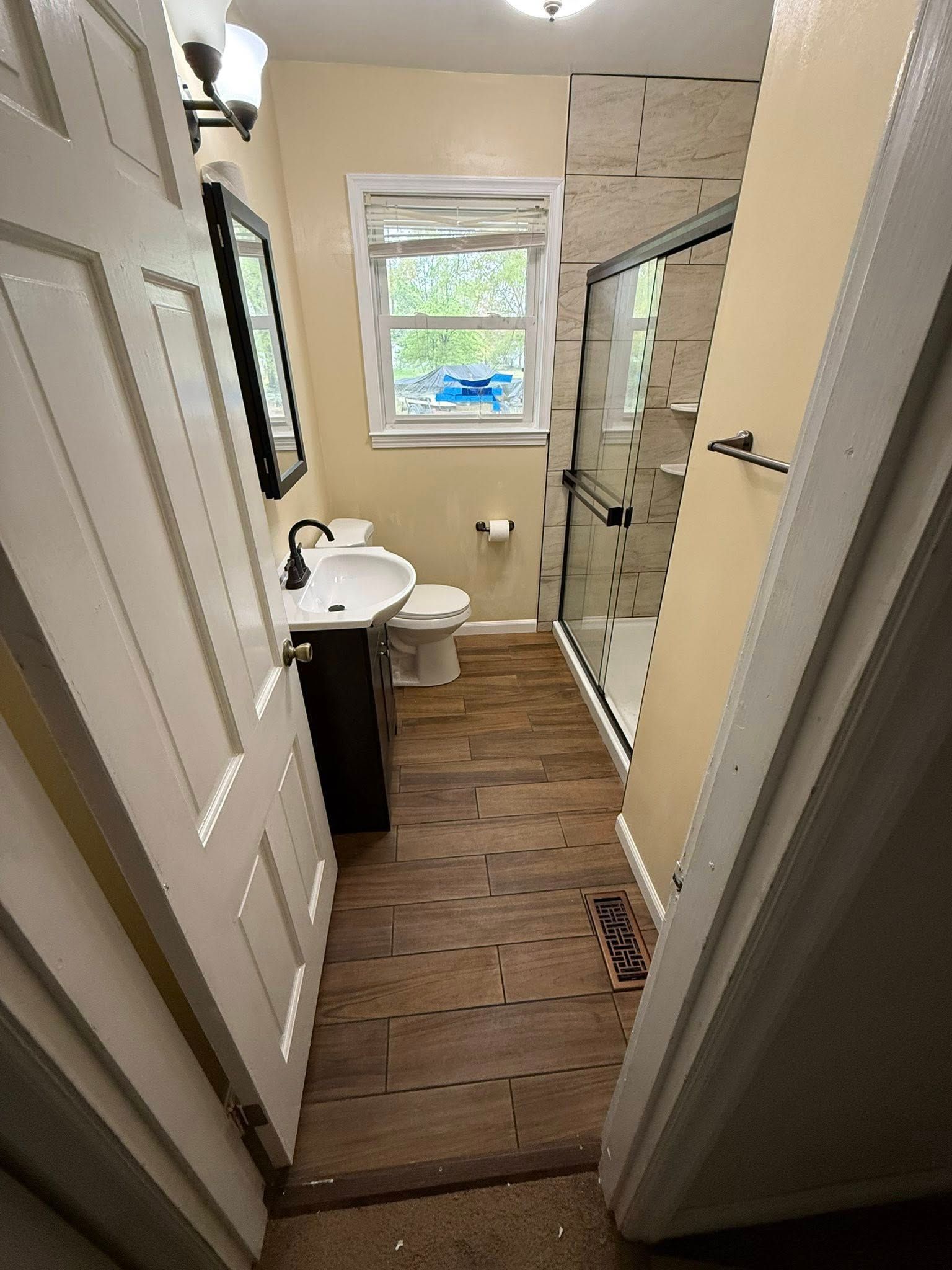 Bathroom with a dark-vanity sink, toilet, glass shower, and wood-look flooring. Light yellow walls and white trim.
