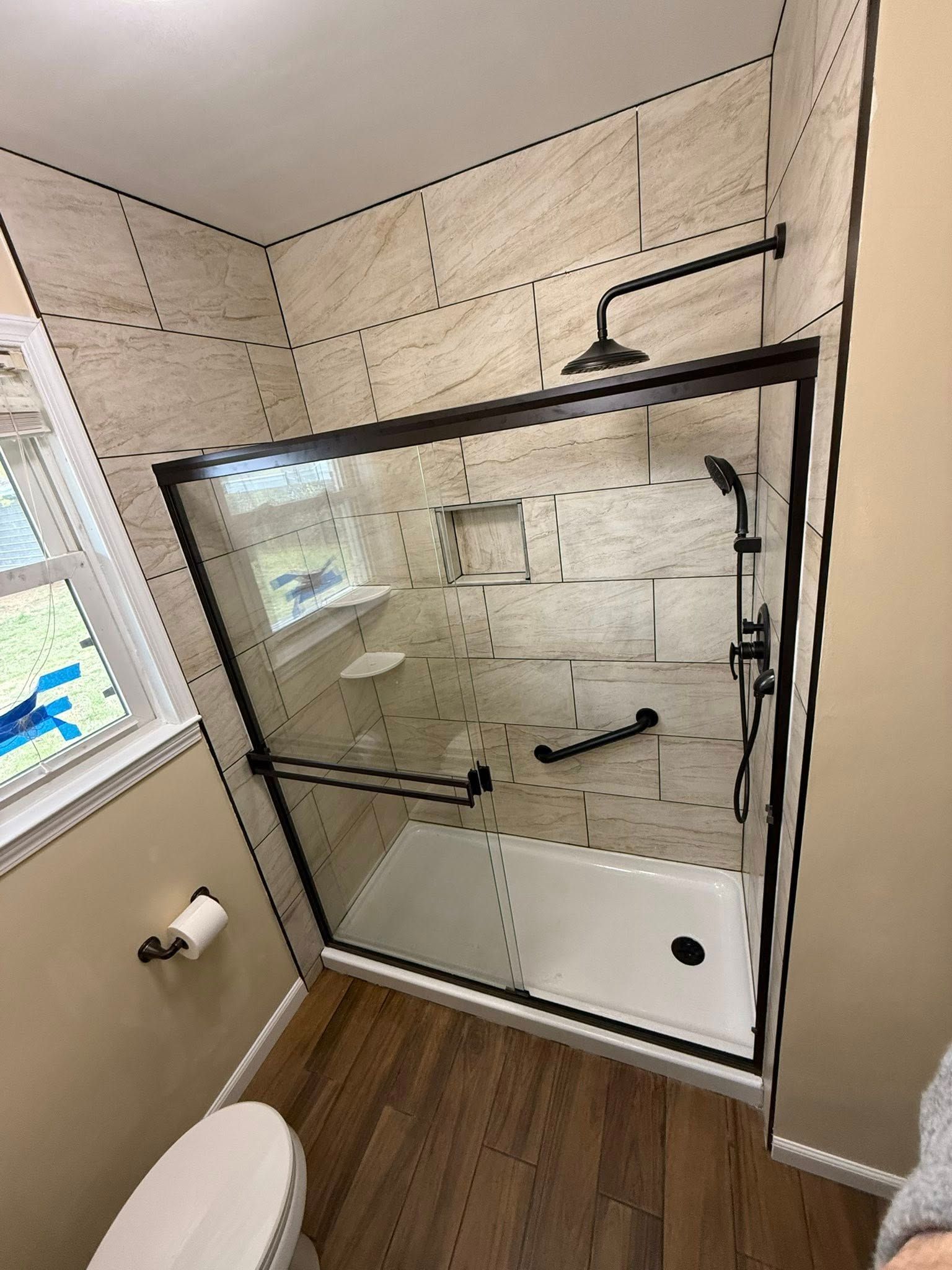 Bathroom with a black-framed glass shower enclosure, beige tiled walls, and wood-look flooring.