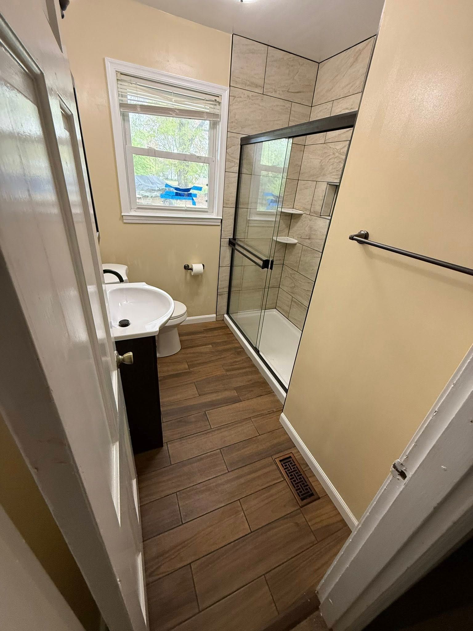 Small bathroom with shower, toilet, and sink. Brown floor, beige walls, and a window.