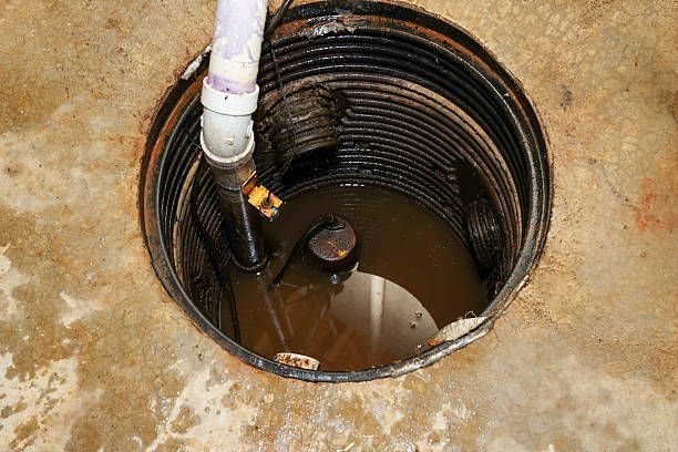A sump pump pit filled with murky water, with a black corrugated wall, PVC pipe and pump inside a concrete floor.