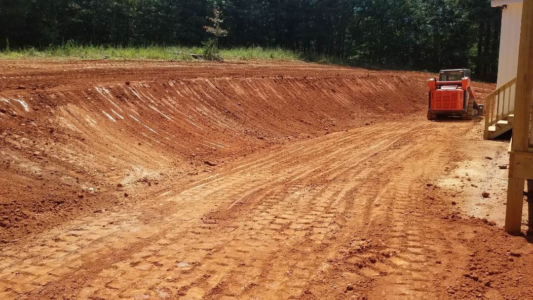 Skid steer grading a dirt construction site. Red dirt, cut bank, and trees in the background.