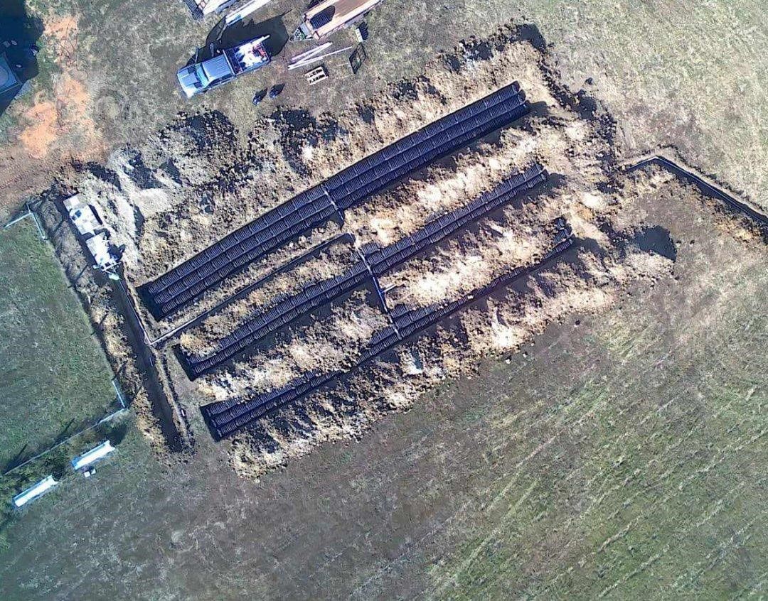 Overhead view of a construction site with black pipes laid in trenches, surrounded by dirt and green field.