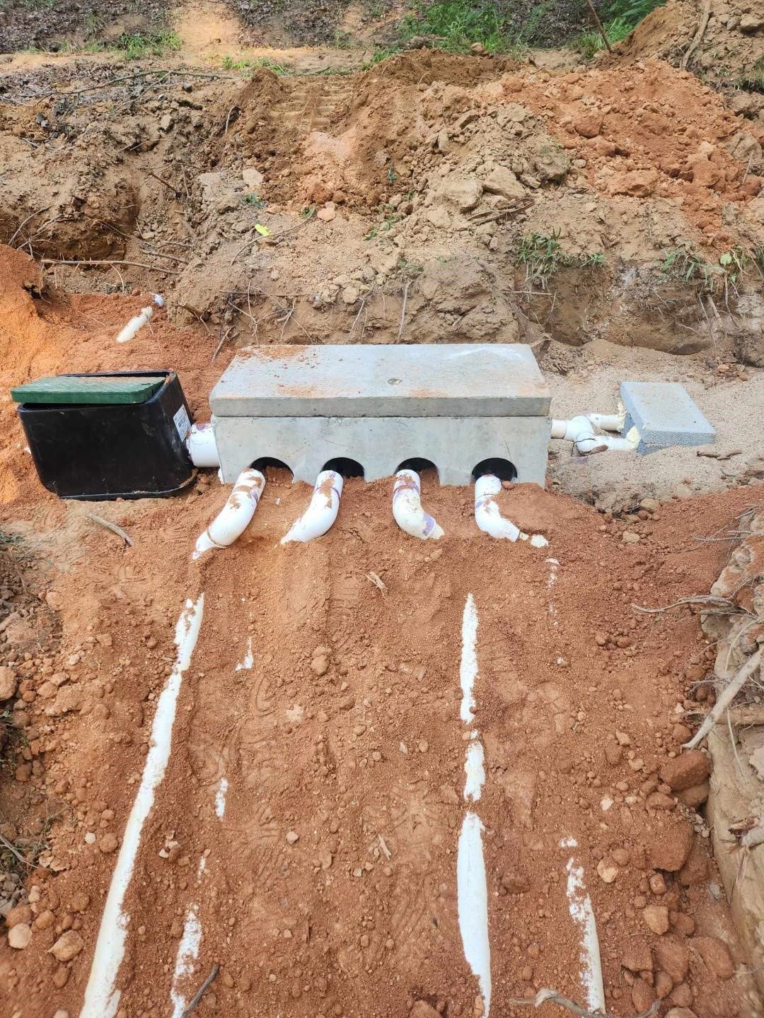 Septic system installation; concrete distribution box with four white pipes leading to trenches in the brown earth.