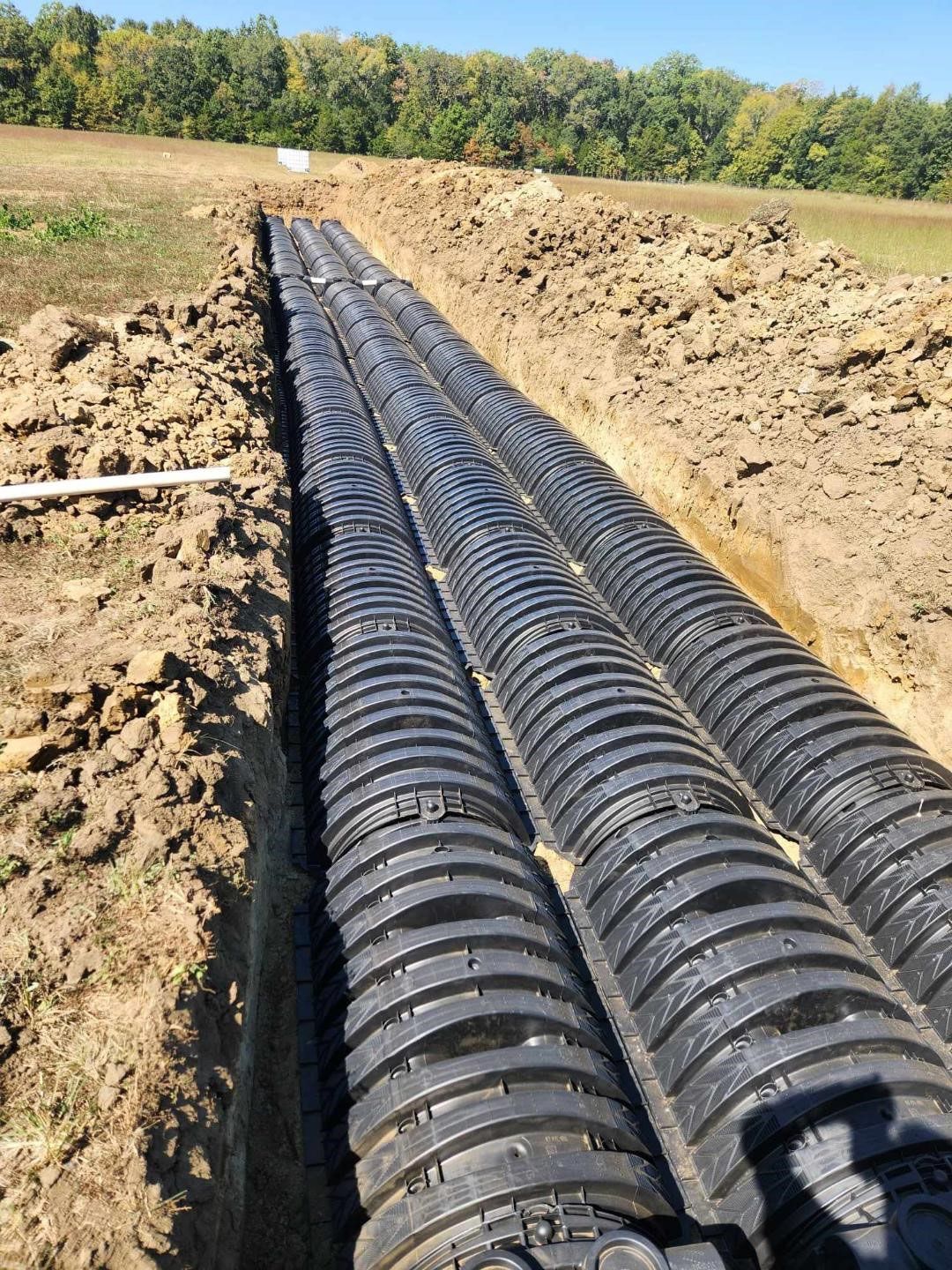 Black corrugated drainage pipes laid in a trench in a field. Brown earth and green trees in the background.