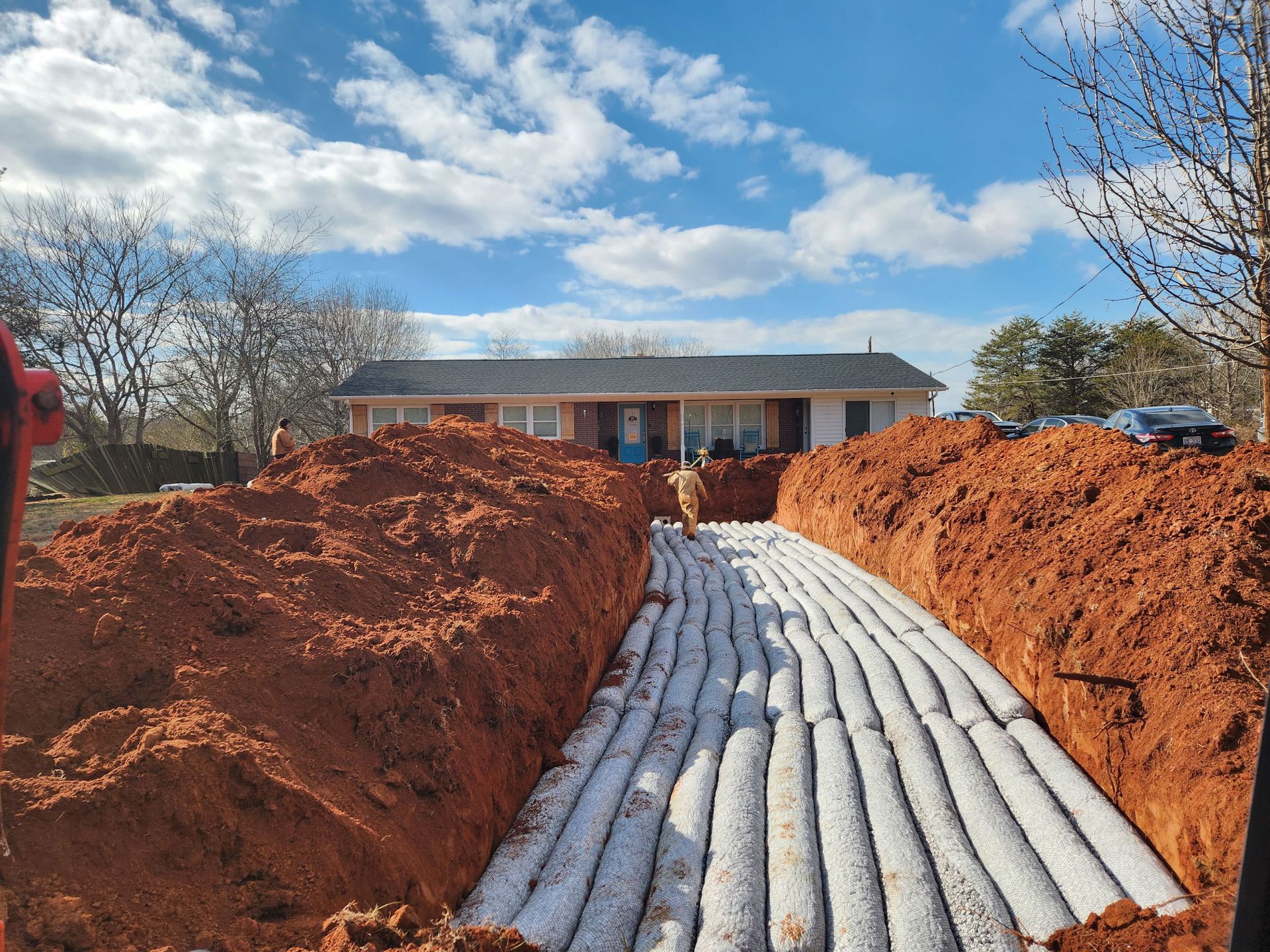Long, excavated trench with white drain field material, in front of a house, under a blue sky.