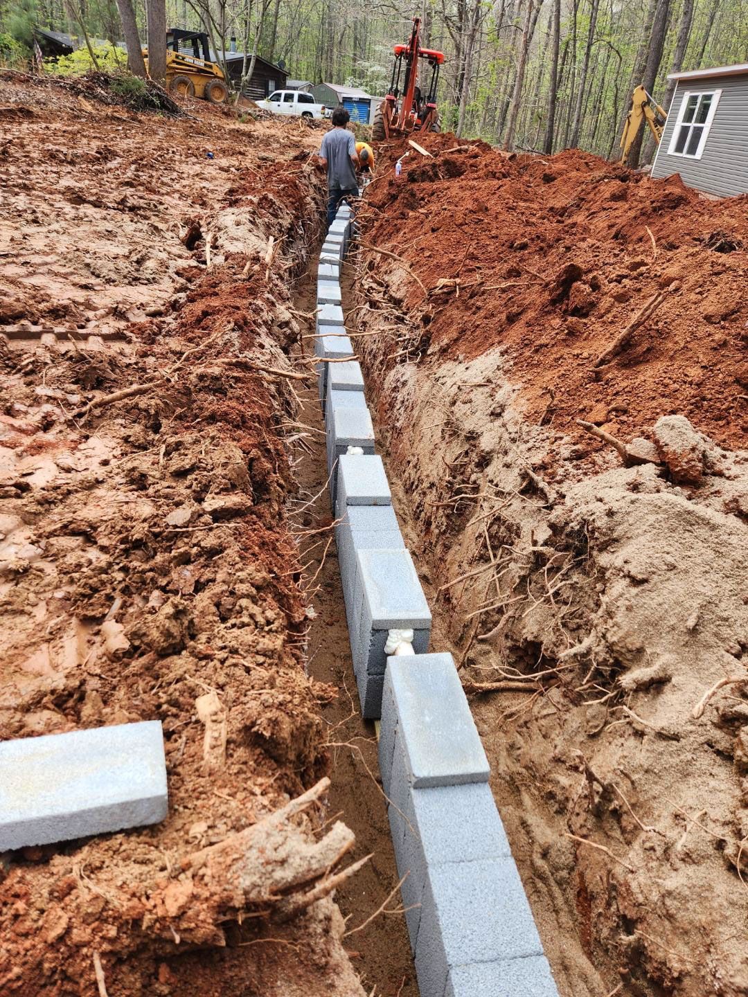 A trench with stacked gray retaining wall blocks, a person walks along it. Red dirt surrounds.