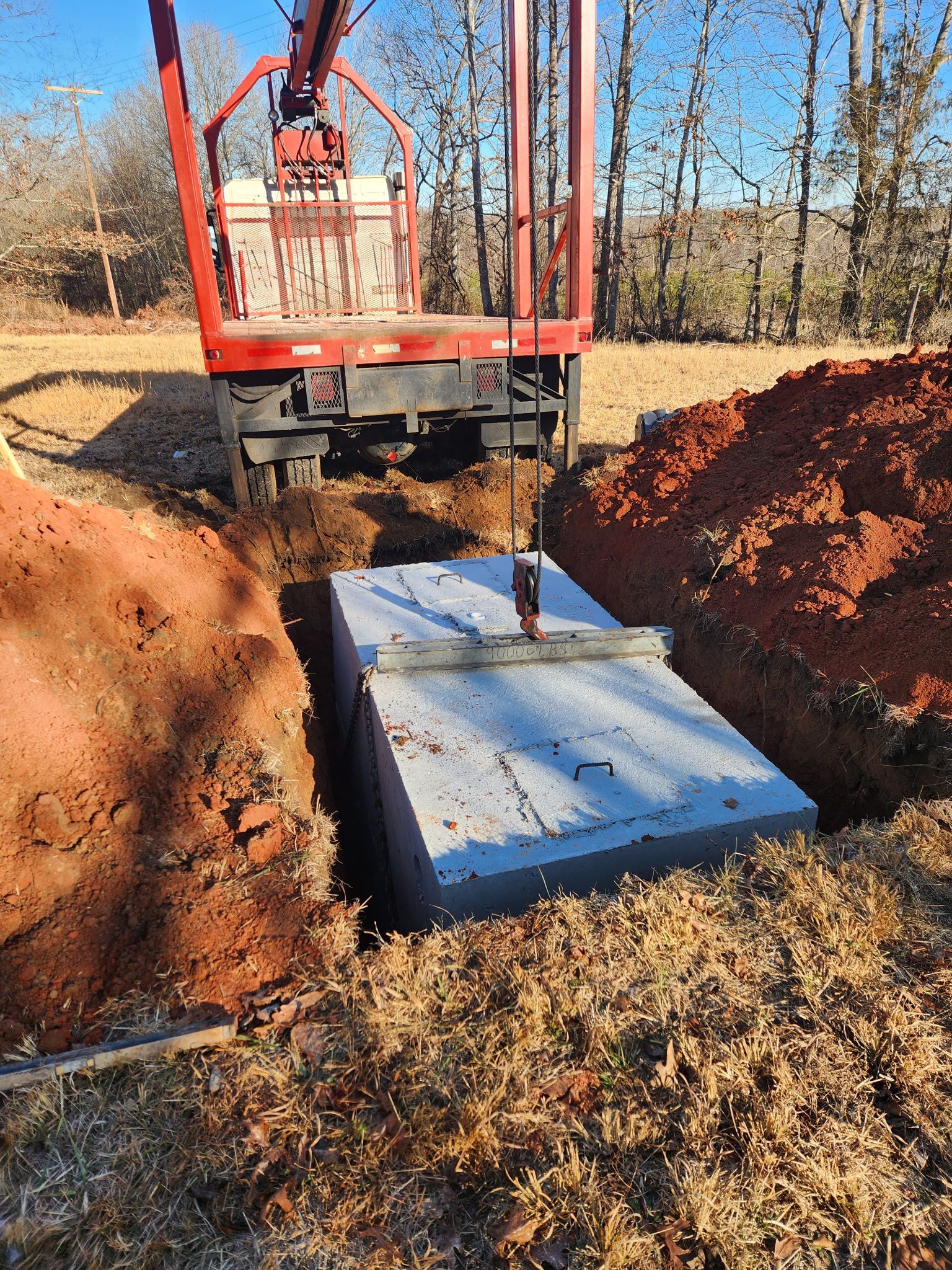 A concrete septic tank is lowered into a hole by a red truck with a crane; outdoor setting.